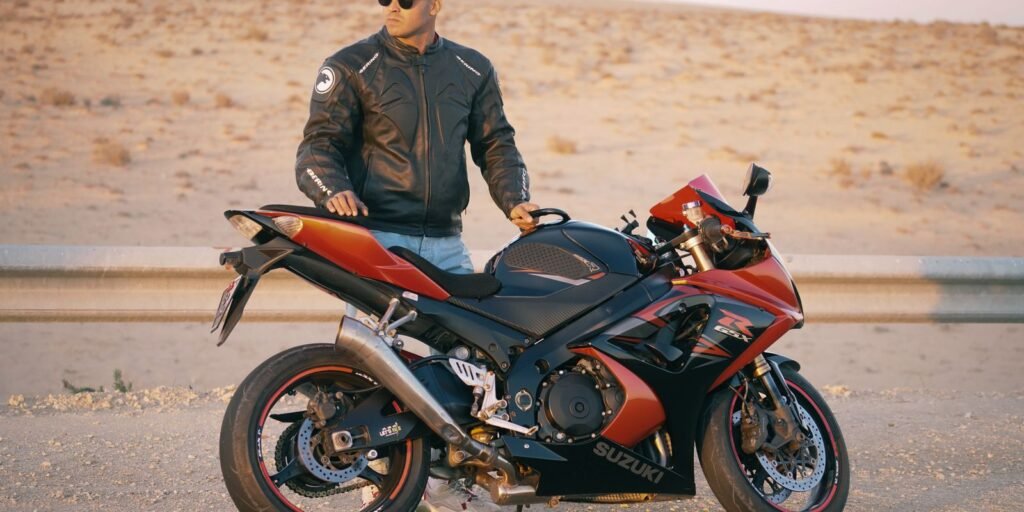 Uomo con giacca di pelle e cappellino in posa con una moto sportiva su una strada nel deserto