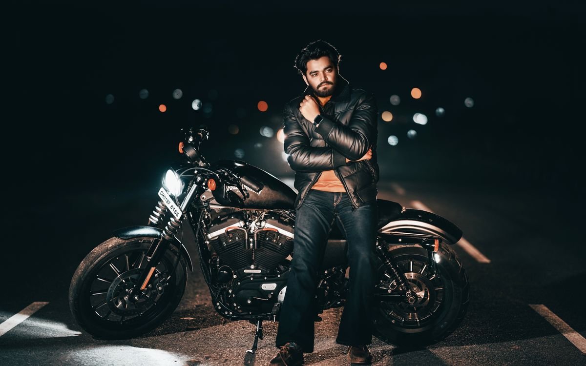 Uomo con giacca di pelle in posa con una moto di notte, sullo sfondo le luci della città