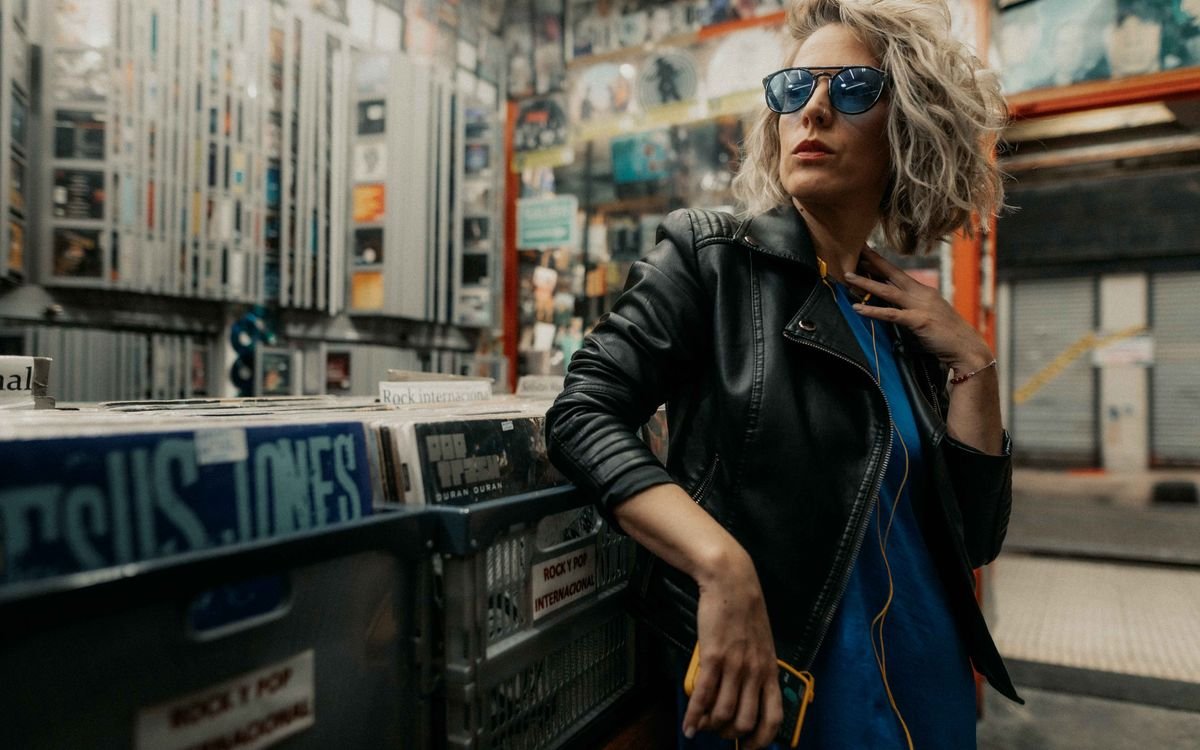 Donna elegante con giubbotto di pelle e occhiali da sole in posa in un negozio di dischi in vinile