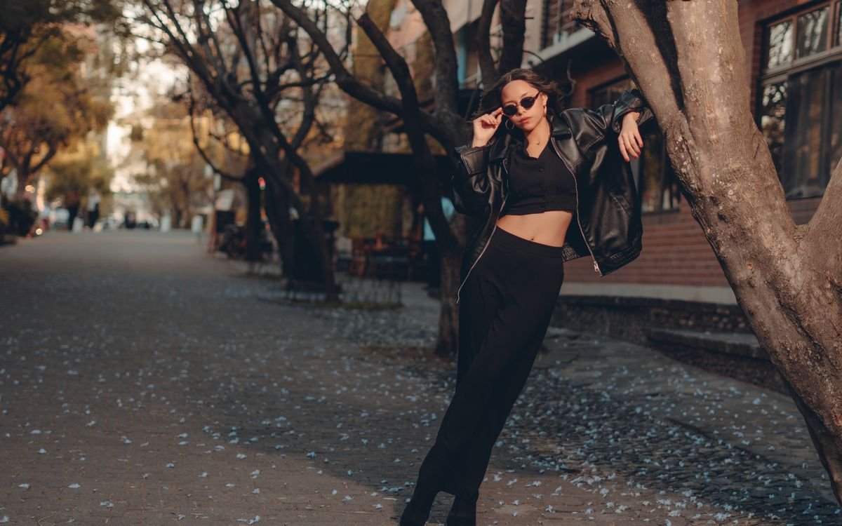 Donna elegante in outfit nero in posa in una scena urbana autunnale con foglie sparse