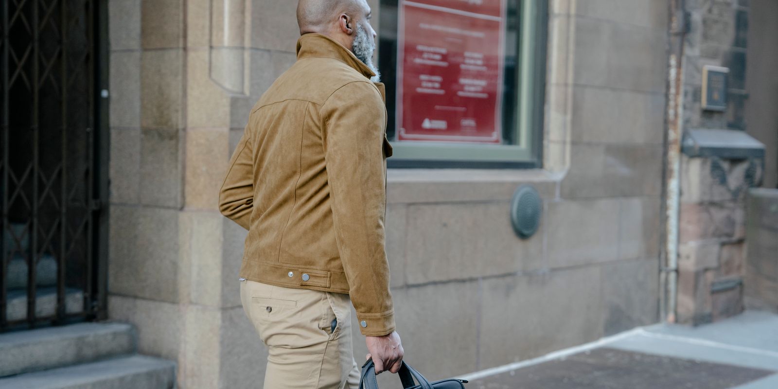 Un uomo con la barba cammina per una strada cittadina portando una valigetta, con indosso una giacca beige