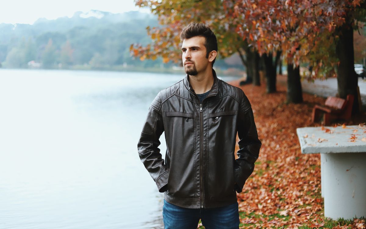 Un uomo con la barba in giacca di pelle marrone in riva a un lago in autunno