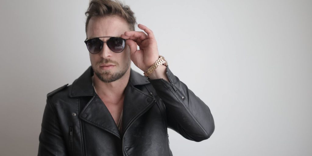 Uomo elegante con giacca di pelle nera e occhiali da sole in posa in interni