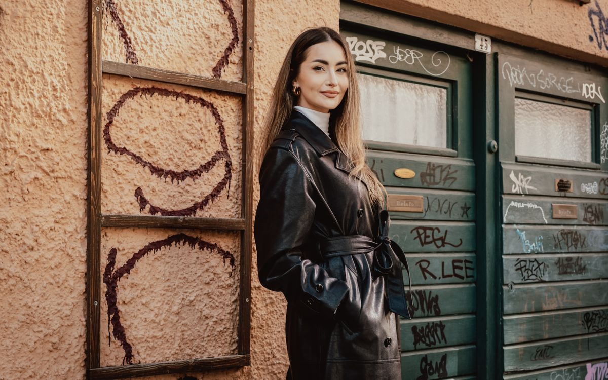 Una donna posa con sicurezza in un lungo cappotto di pelle nero davanti a un muro urbano