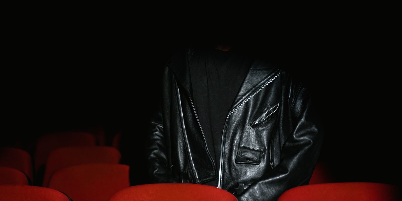 Una persona con giacca di pelle nera seduta in un cinema in penombra tra poltrone rosse