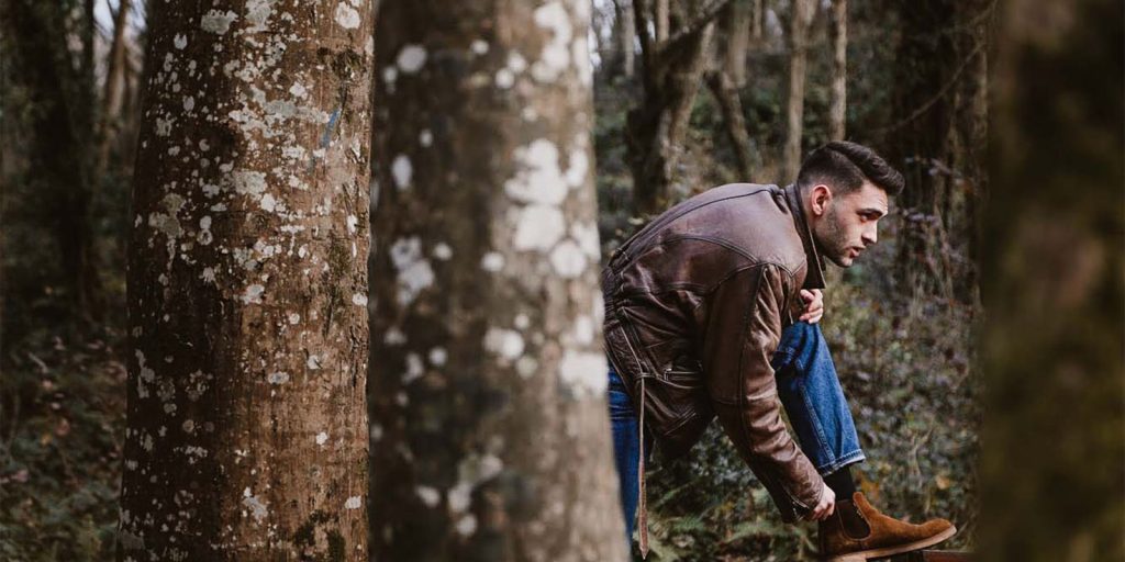 Un uomo con giacca di pelle marrone allaccia stivali di pelle marrone su un sentiero nel bosco