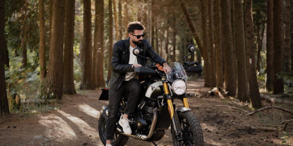Un uomo con una giacca di pelle marrone si ferma con una moto su un tranquillo sentiero nel bosco